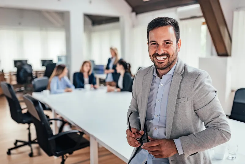 Homem sorridente em um terno está de pé na frente de uma mesa de conferência